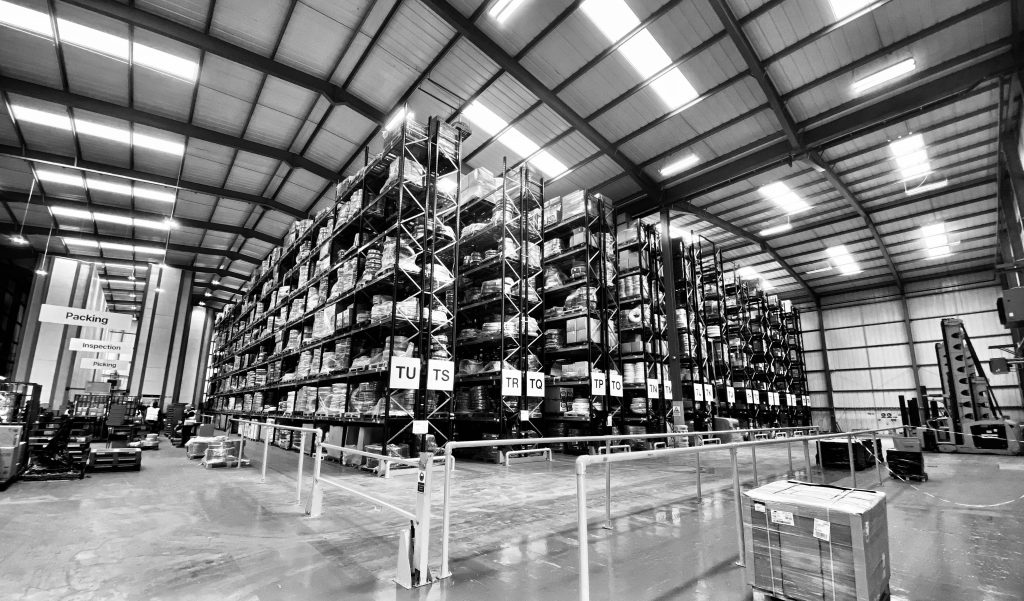 Black and white wide area shot of a warehouse showing tall racking and signs for various work areas