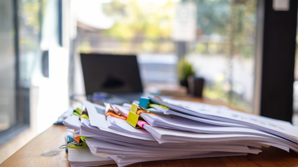 Documents held together by bulldog clips in a large stack on a desk